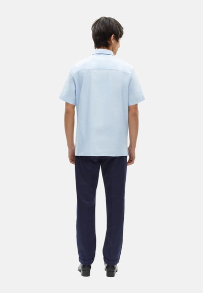 Camisa de manga corta azul claro fabricada en un tejido transpirable, con un corte recto y una costura trasera en contraste, combinada con pantalones oscuros en color navy.