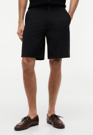 Mężczyzna w czarnych szortach do kolan i brązowych skórzanych mokasynach zapinanych na sznurówki, stojący z rękami w kieszeniach na białym tle.