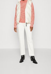 Bílá péřová vesta přes červený pletený svetr, doplněná o bílé zkrácené kalhoty a černé vyšší boty na podpatku. Hladké materiály a moderní designové detaily.