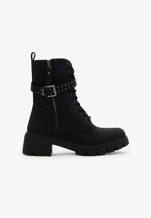 Botín negro hasta el tobillo con tacón grueso, cremallera lateral, parte delantera con cordones y detalle de correa con hebilla con tachuelas sobre fondo blanco.