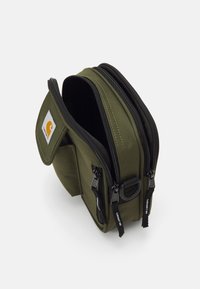 Kompakt olivgrøn Carhartt pouch med to lynlåslommer og en frontlomme med klap, vist let åben på hvid baggrund.