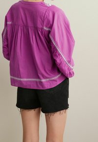Lilla bomuldsbluse med pufærmer, hvide broderede accenter, samlet ryg, parret med sorte denimshorts med frynset kant.