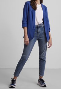 Blazer bleu cintré sur un t-shirt blanc, associé à un jean clair et des chaussures de sport multicolores. Design simple et structuré.