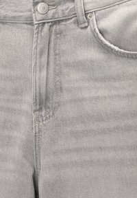 Gros plan sur un jean en denim gris clair montrant la ceinture, le bouton, le passant de ceinture, la poche avant et les coutures visibles.