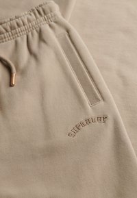 Pantaloni della tuta beige con vita elasticizzata, dotati di una tasca e logo "SUPERDRY" in rilievo di colore complementare. Tessuto morbido e strutturato.