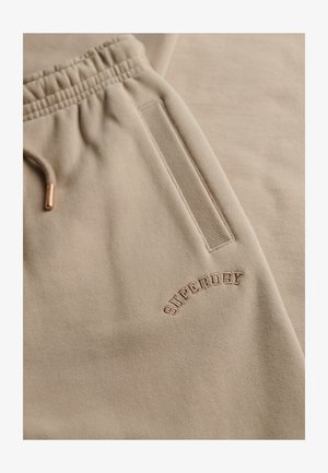 Beige sweatpants med elastisk talje, med en lomme og prægede "SUPERDRY" logo i komplementær farve. Blødt, tekstureret stof.