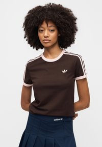 T-shirt en coton marron avec des accents roses et trois bandes roses sur les épaules. Présente un petit logo blanc sur la poitrine et un col rond.