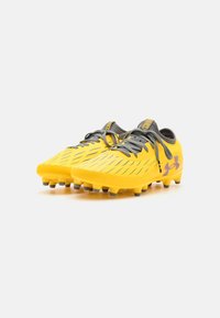 Sárga focicipők texturált felsőrésszel, szürke fűzőkkel és formázott tüskékkel. Az Under Armour logóját és kontrasztos díszítéseket tartalmaz.