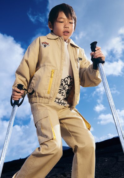 Veste beige avec des accents jaunes, arborant un graphique de lion sur une chemise blanche et un pantalon beige assorti, tenant des béquilles sur un fond de ciel bleu.