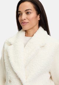 Hvid faux fur frakke med bred krave og dobbeltknaplukning. Strikkede detaljer på siden og en subtil guldkknaptilføjelse giver tekstur.