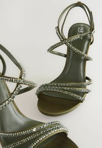 Sandali verdi con cinturini, dettagli metallici e ornamenti in strass, dotati di tacco alto e una texture liscia e opaca.