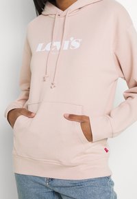Ljusrosa hoodie i mjukt tyg, med en framficka och vit Levi's-logotyp. Dragsnören vid huvan, med en röd etikett vid fållen.