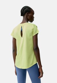 Citrānas zaļas krāsas īsām piedurknēm blūze ar izgriezumu aizmugurē, apvienota ar zilu pieguļošu biksēm. Gluda tekstūra un brīvs piegriezums.