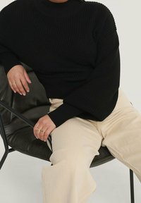 Czarny dzianinowy sweter z wysokim kołnierzem i szerokimi rękawami, w połączeniu z jasnobeżowymi spodniami typu wide-leg. Osoba nosi pierścionki na dłoniach.