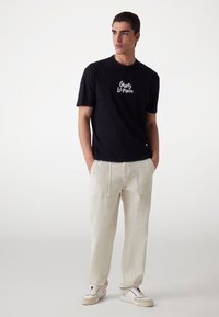 Camiseta negra de manga corta con el logo blanco "Guess Los Angeles", combinada con pantalones de algodón beige claro de pierna ancha y zapatillas blancas.