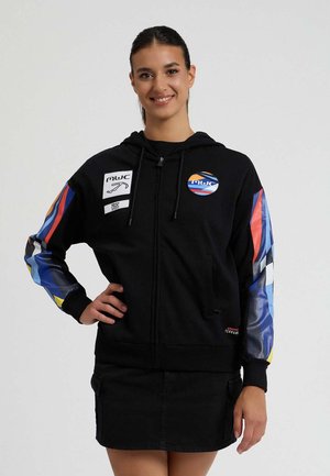 Felpa nera con zip e maniche a fantasia colorata, caratterizzata da un logo circolare e toppe. Realizzata in tessuto morbido con cappuccio regolabile.