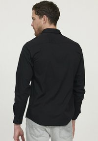 Chemise noire à manches longues au toucher lisse, dotée d'un col, de poignets boutonnés et d'un ourlet arrondi. La vue arrière met en avant les détails de couture.