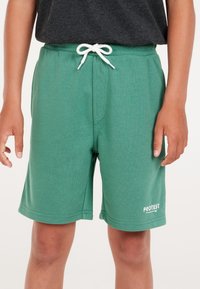 Groene katoenen shorts met een elastische tailleband en witte trekkoorden, voorzien van een witte "PROTEST" branding aan de onderkant vooraan.