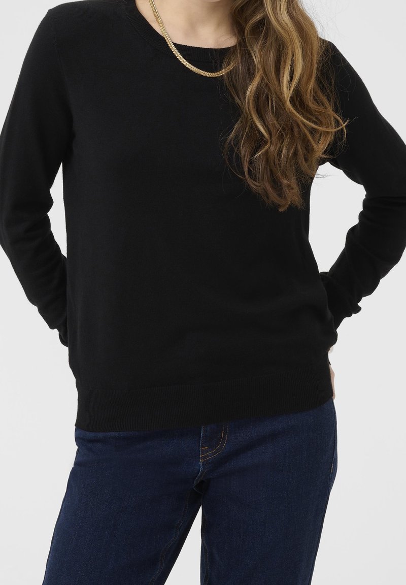 Femme aux longs cheveux ondulés portant un pull noir, un collier en or et un jean bleu, debout les mains dans le dos.