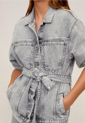 Vrouw draagt een lichtgrijze acid-wash spijkerjas met voorzakken, knoopsluiting en een ceintuur die is gestrikt, handen in de onderste zakken.