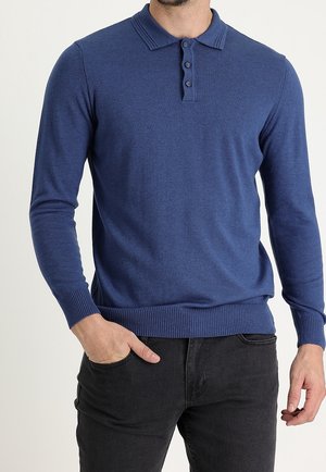 Mężczyzna ubrany w niebieski sweter polo z długim rękawem, z prążkowanymi mankietami i dołem, do tego czarne jeansy, ręka w kieszeni.