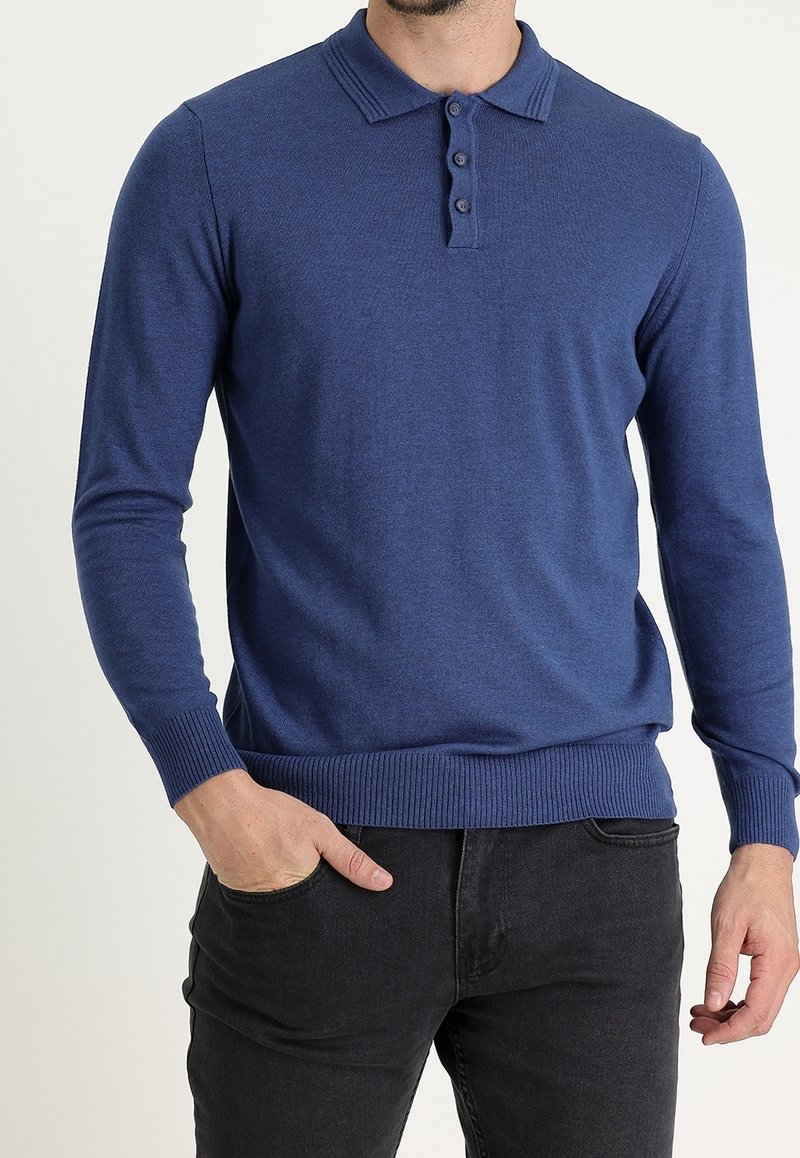 Homme portant un pull polo bleu à manches longues avec poignets et ourlet côtelés, associé à un jean noir, main dans la poche.