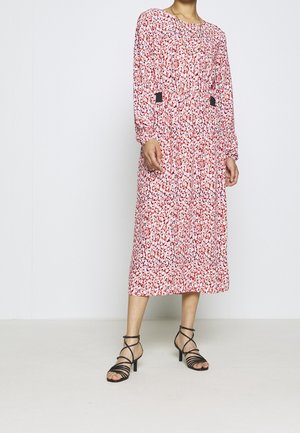 Femme portant une robe midi rose à fleurs à manches longues avec des accents noirs à la taille, associée à des sandales à talons noires à brides.