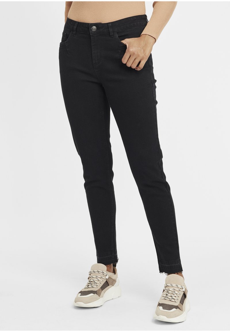 Jean skinny en denim noir avec fermeture par bouton, poches avant et ourlet effiloché, associés à des baskets athlétiques beige et blanches.