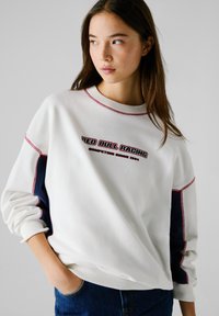 Felpa bianca con dettagli laterali blu navy e cuciture rosse. Presenta una grafica "RED BULL RACING" sulla parte anteriore, scollatura rotonda e vestibilità rilassata.