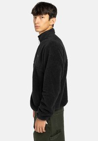 Pullover in pile nero con colletto alto, tasca frontale con zip e maniche lunghe. Indossato sopra pantaloni verdi, in posizione di profilo.