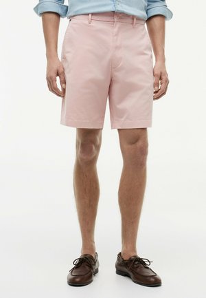 Mann trägt hellrosa Shorts, hellblaues Hemd mit hochgekrempelten Ärmeln und braune Leder-Schnürschuhe, steht vor einem schlichten weißen Hintergrund.