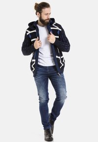 Strick-Navy-Cardigan mit weißen und blauen geometrischen Mustern, ausgestattet mit einer Kapuze, einem Reißverschluss vorne und strukturierten Ärmeln. Getragen mit verwaschenen Jeans und schwarzen Stiefeln.