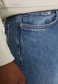 Niebieskie dżinsowe spodnie z teksturowanym wykończeniem, z kieszenią na monety, widocznymi szwami i lekkim zatarciem. Ręka widoczna na pasku.