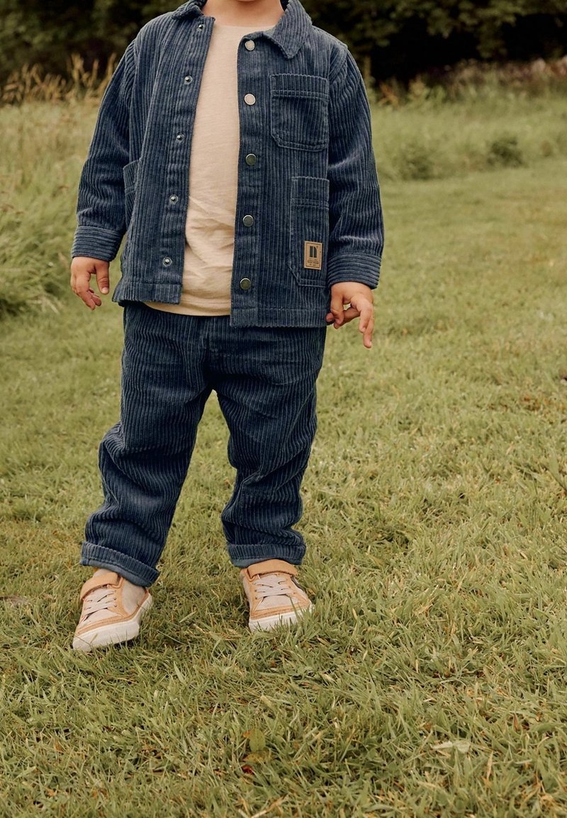 Tout-petit debout sur l'herbe portant une veste et un pantalon en velours côtelé bleu avec les revers roulés, une chemise beige et des baskets beiges.