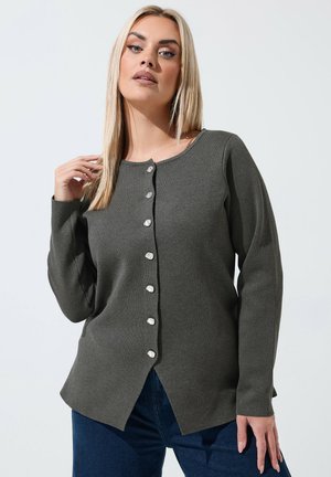 Kvinde med langt blondt hår iført en mørkegrå knapcardigan og blå jeans, poserer mod en ensfarvet lys baggrund.