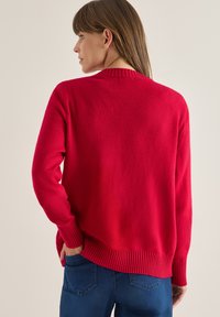Suéter de punto rojo con cuello y puños acanalados. El diseño presenta un corte holgado y una parte trasera ligeramente más larga. Textura suave visible.