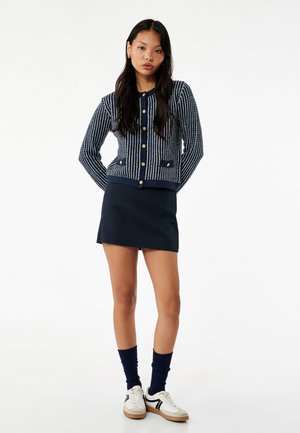 Marineblauwe gebreide cardigan met verticale witte strepen, gouden knopen en twee zakken; gecombineerd met een marineblauwe mini-jurk en witte sneakers.