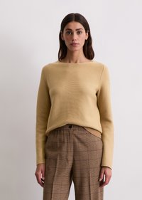Beżowy sweter dziewiarski z szerokim dekoltem i długimi rękawami, zestawiony z brązowymi spodniami w kratę o luźnym kroju i z detalami w postaci guzików.