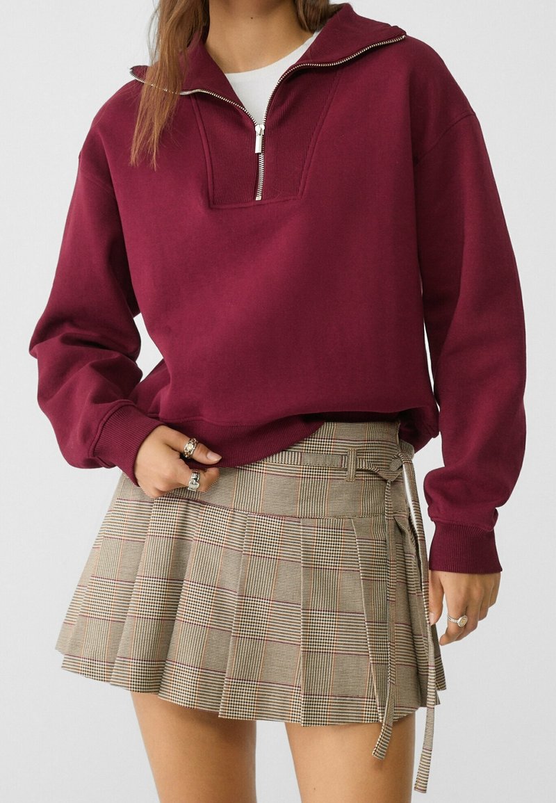 Femme portant un sweat-shirt bordeaux à fermeture éclair sur le devant et une jupe plissée beige à carreaux avec un détail ceinture.