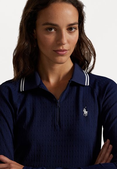 Navy strikket zip-up polo med hvite aksentstriper på kragen, med et teksturert kabeldesign og en liten hvit logo på brystet.