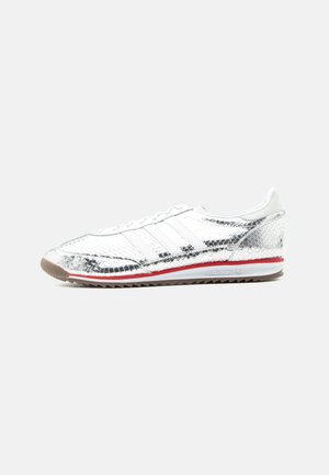 Sølvfarget metallisk teksturert lav høyde sneaker med hvite lisser, rød stripe på hvit såle og rillestøpt brun yttersåle.