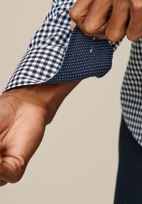 Poignet de chemise à carreaux bleu et blanc montrant un tissu intérieur contrastant bleu marine avec de petits pois blancs. Le poignet a une finition soignée et des coutures nettes.