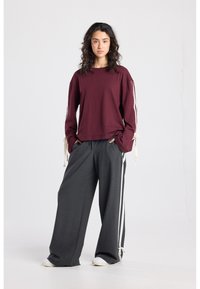 Donkerrode top met lange mouwen en strikjes bij de manchetten, gecombineerd met grijze broek met wijde pijpen en witte zijstrepen, afgewerkt met witte sneakers.