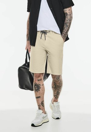 Beige shorts med snoretræk i taljen, parret med en sort skjorte med knapper og hvide sneakers. Sort taske ved siden af.