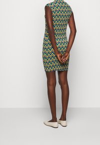 Bunt gemustertes Kleid mit Zickzack-Design in Grün, Blau und Gelb, in taillierter Form und ohne Ärmel, kombiniert mit hellen flachen Schuhen.