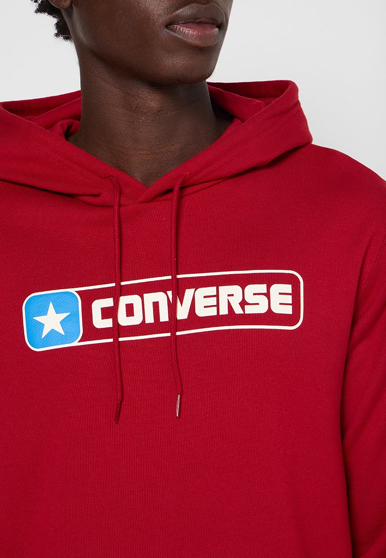 Osoba ubrana w czerwoną bluzę z kapturem, na której widnieje białe i niebieskie logo Converse, ukazująca sznurki oraz częściowy widok szyi i warg.