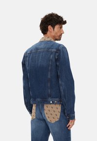 Chaqueta de mezclilla en azul oscuro con un corte clásico, que presenta cierre de botones y costuras decorativas; el interior tiene un forro con un patrón.