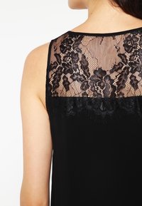 Svart ärmlös topp med en bakpanel i spets med blommönster och vågiga kanter, slät tyg med en lätt lyster.