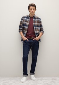 Camicia a quadri marrone, blu e bianca sopra una t-shirt bordeaux, abbinata a jeans blu scuro e sneakers bianche. Jeans con orlo risvoltato.