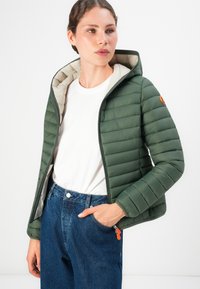 Vrouw in een groene gewatteerde hoodie-jack met oranje ritssluitingen, een wit T-shirt en blauwe high-waisted jeans, poserend met één hand in de zak.